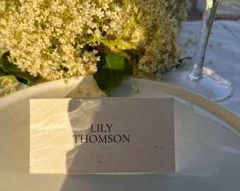 Könnte beinhalten: Eine Tischkarte mit dem Namen "LILY THOMSON" in Schwarz auf einer strukturierten, cremefarbenen Karte gedruckt. Die Karte liegt auf einem weißen Teller, mit einem Glas Champagner und einem Strauß weißer Blumen im Hintergrund.