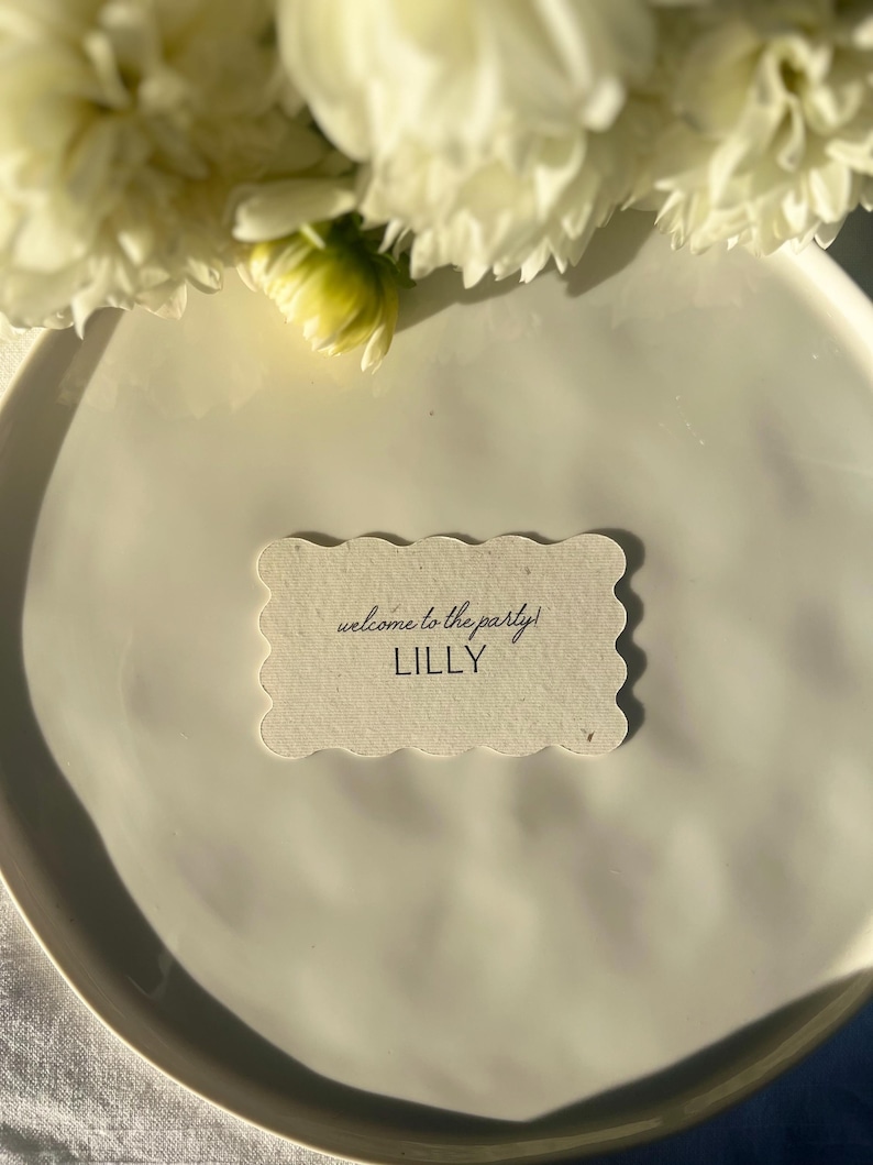 K&ouml;nnte beinhalten: Eine wei&szlig;e Platzkarte mit gewelltem Rand und dem Text "welcome to the party!" sowie dem Namen "LILLY" in schwarzer Schrift. Die Karte liegt auf einem wei&szlig;en Teller, mit wei&szlig;en Blumen im Hintergrund.