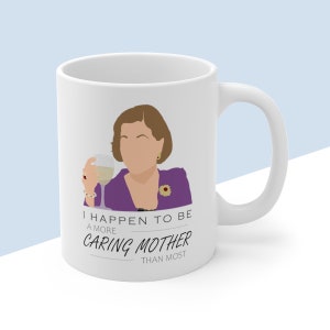 Può includere: Una tazza in ceramica bianca con un'illustrazione viola e nera di una donna che tiene un bicchiere di vino. Il testo sulla tazza recita "I happen to be a more caring mother than most."