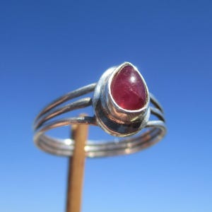 Puede incluir: Anillo de plata con una piedra preciosa roja oscura en forma de lágrima. El anillo tiene un diseño de triple banda y se muestra contra un cielo azul claro. La piedra preciosa está engastada en un bisel de plata.