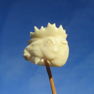 May include: A carved ivory-coloured moose head on a wooden stick, set against a clear blue sky. The moose has detailed features, including antlers and a defined snout. The carving is held by a thin, brown stick.