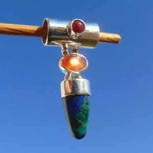 Op de afbeelding: Een zilveren hanger met een rode edelsteen, een oranje edelsteen en een blauwgroene edelsteen. De hanger hangt aan een houten deuvel tegen een helderblauwe lucht.