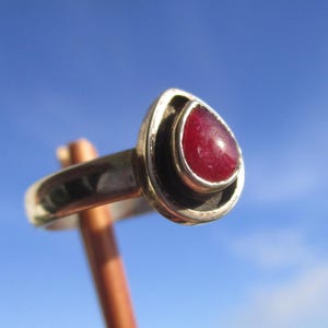 Könnte beinhalten: Ein Silberring mit einem tropfenförmigen roten Edelstein, der in einer glatten Metallfassung sitzt. Der Ringreif ist aus poliertem Silber und die Fassung weist einen dunklen, vertieften Bereich um den zentralen roten Stein auf.