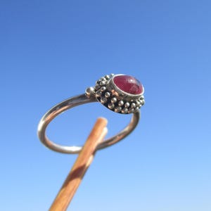 Könnte beinhalten: Ein silberner Ring mit einem großen, ovalen, roten Edelstein. Die Fassung des Rings ist mit kleinen, strukturierten Details verziert. Der Ring wird vor einem klaren blauen Himmel gehalten.