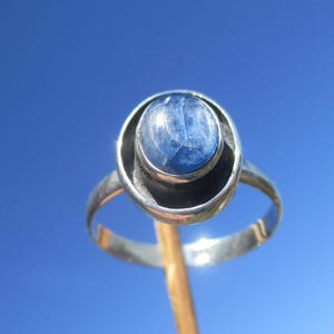 Op de afbeelding: Een zilveren ring met een grote, ovale, blauwe edelsteen gezet in een cirkelvormige bezel. De band van de ring heeft een eenvoudig, gebogen ontwerp. De achtergrond is een heldere, lichtblauwe lucht, die de details van de ring benadrukt.