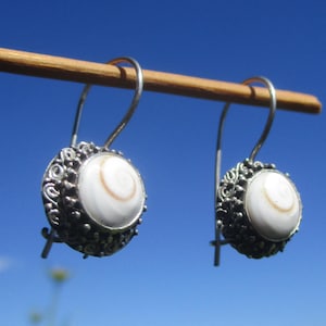 Op de afbeelding: Een paar zilveren oorbellen met ronde, witte Shiva-oog schelp cabochons gezet in gedetailleerde, donker geoxideerde zilveren filigraan monturen met gebogen oorhaken, hangend aan een dunne houten stok tegen een helderblauwe lucht.
