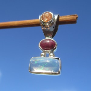 Può includere: Un pendente in argento con tre pietre preziose: una pietra solare rotonda arancione, un rubino ovale marrone e un'opale rettangolare con lampi blu e verdi. Il pendente è sospeso contro un cielo blu chiaro.