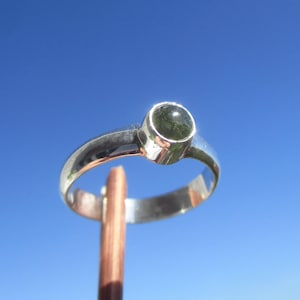 Puede incluir: Un anillo de plata con una gema redonda de color verde oscuro engastada en un bisel. La banda del anillo, pulida y reflectante, presenta un diseño sencillo y elegante. El anillo se muestra contra un cielo azul brillante.
