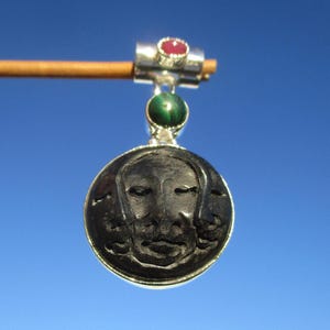 May include: A silver pendant featuring a carved black face design. The pendant is suspended from a silver bar with a red gemstone and a green gemstone. Set against a clear blue sky.