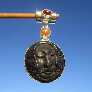 May include: A silver pendant featuring a carved black face and two gemstones. The top gemstone is red and the bottom gemstone is orange.
