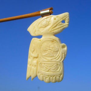 Puede incluir: Colgante tallado de color marfil que representa una figura estilizada de ave o animal. El colgante presenta detalles intrincados, incluyendo alas talladas, una cara y otros elementos simbólicos. Un palo de madera está unido a la parte superior con un anillo de metal.