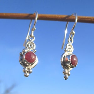 Op de afbeelding: Zilveren oorbellen met een rode edelsteen. De oorbellen hebben een spiraalvormig ontwerp boven de edelsteen en een cluster van kleine zilveren kralen eronder. Helderblauwe lucht.