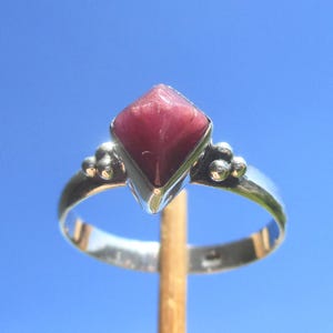 Könnte beinhalten: Ein Silberring mit einem zentralen, pyramidenförmigen, dunkelrosa Edelstein. Die Ringschiene besteht aus poliertem Silber und ist auf beiden Seiten der Steinfassung mit dekorativen Gruppen aus drei kleinen Silberkugeln versehen, was für Symmetrie sorgt.
