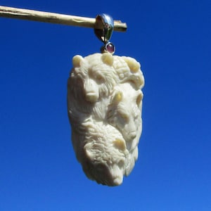 May include: A cream-coloured pendant carved into the shape of three bear heads stacked vertically. The pendant features a silver-toned metal bail at the top, accented with a small, round, dark red gemstone set directly below the bail attachment point.