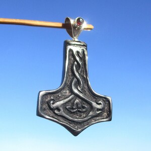 Può includere: Un ciondolo color argento a forma di martello di Thor, con intricati disegni a nodo celtico. Il ciondolo ha una piccola gemma rossa in cima ed è sospeso da un anello d'argento. Lo sfondo è un cielo blu chiaro.