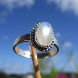 Op de afbeelding: Zilveren ring met een ovale, iriserende maansteen. De maansteen is gezet in een kralenrand. De ring wordt getoond tegen een blauwe lucht.