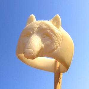 May include: A cream-coloured ring featuring a detailed, three-dimensional carving of a wolf head on the front. The wolf design includes textured fur lines, pointed ears, and a defined snout, all integrated into the smooth, solid band of the ring.