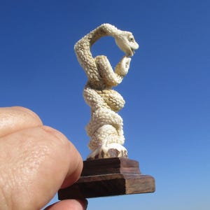 May include: A small, off-white, textured snake figurine coiled vertically, with its head curved toward its tail. The sculpture is mounted on a dark brown, square wooden base, showcasing intricate scale details across the entire body of the snake.