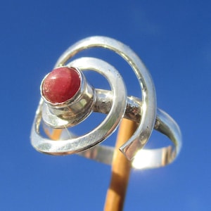 Op de afbeelding: Een zilveren ring met een gepolijste, ronde, dieprode edelsteen in een zetting. De band is ontworpen met een opvallende, open spiraalvormige metalen structuur die de centrale steen omringt, wat zorgt voor een gelaagd en wervelend uiterlijk in het metaal.