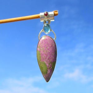 Puede incluir: Un colgante en forma de lágrima con un engaste de plata. El colgante presenta una piedra grande y pulida con una mezcla de tonos morados y verdes. Una pequeña piedra roja redonda y una piedra ovalada azul están engastadas encima de la piedra principal.