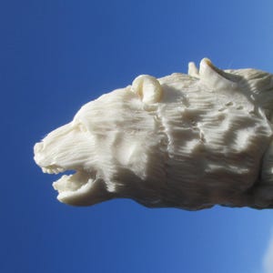 Op de afbeelding: Een gedetailleerde, crèmekleurige sculptuur van een berenkop tegen een helderblauwe lucht. De kenmerken van de beer zijn ingewikkeld gesneden, met zichtbare vachttextuur en een open mond. De sculptuur is een close-up, die het vakmanschap benadrukt.
