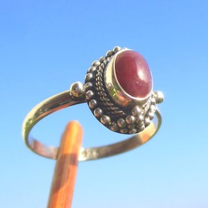 Könnte beinhalten: Ein Silberring mit einem glatten, ovalen, dunkelroten Edelstein in einer Fassung. Die Fassung ist mit einem gedrehten Seilmuster und kleinen Silberkugeln verziert. Der Ring besteht aus poliertem Silber und weist einen sichtbaren 925er Stempel auf.