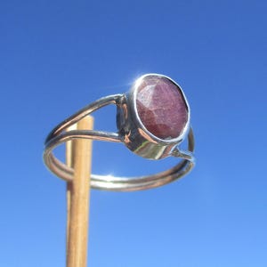 Könnte beinhalten: Ein silberner Ring mit einem großen, oval geformten, facettierten, tiefroten Edelstein. Der Ring hat ein geteiltes Schaftdesign mit zwei Bändern, die sich um den Finger krümmen. Der Edelstein ist in einer Lünettenfassung gefasst.