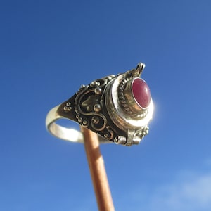 Könnte beinhalten: Ein silberfarbener Ring mit einem ovalen, dunkelrosa Cabochon-Stein, der in eine gedrehte Metallfassung eingefasst ist. Die Ringschiene und die Fassung sind mit filigranen Metallverzierungen, kleinen Kugeln und Blattmotiven versehen.