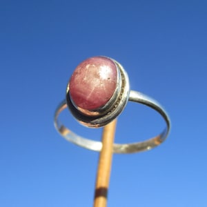 Può includere: Un anello d'argento con una grande gemma rosa di forma ovale. La gemma è incastonata in una lunetta con un bordo decorativo. L'anello è tenuto contro un cielo blu brillante, evidenziando il design e il colore.