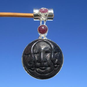May include: A silver pendant with a carved face design, featuring two red gemstones. The pendant is suspended from a silver bail and a wooden stick. The background is a clear, bright blue sky. The pendant is a unique piece of jewellery.