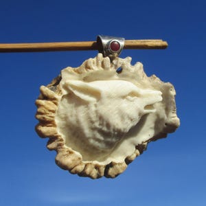 Op de afbeelding: Een hanger met een gebeeldhouwde wolfskop in een getextureerde, gebroken witte geweibasis. Het heeft een zilveren beugel met een kleine, ronde, rode steen. De achtergrond is een heldere, lichtblauwe lucht.