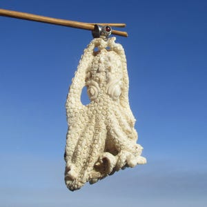 Peut inclure: Pendentif poulpe de couleur ivoire avec une surface texturée, suspendu contre un ciel bleu. Le pendentif est maintenu par une baguette en bois et un anneau argenté avec une gemme rouge. Le design du poulpe présente des tentacules détaillés et une ouverture centrale en forme d'œil.