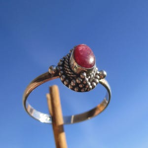 Könnte beinhalten: Ein silberner Ring mit einem großen, ovalen, roten Edelstein. Der Ring hat ein detailliertes Design um den Edelstein herum, mit kleinen Silberperlen und einem gedrehten, seilartigen Rand. Der Ring ist dünn und poliert, vor einem klaren blauen Himmel.