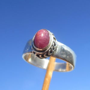 Peut inclure: Bague en argent ornée d'une grosse pierre rose ovale. La pierre est sertie dans une lunette décorative avec un motif ajouré. L'anneau de la bague est lisse et poli. Bijou classique.