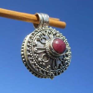 Può includere: Un ciondolo rotondo in argento con una pietra rossa circolare centrale incastonata in un bordo di filo intrecciato. La superficie è decorata con intricati motivi metallici in rilievo e un bordo perlato. Una contromaglia in argento è fissata in alto.