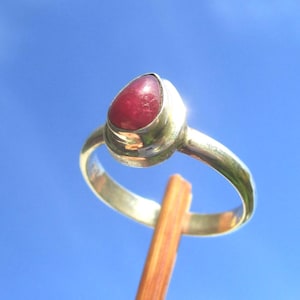Könnte beinhalten: Ein Silberring mit einem tropfenförmigen, tiefroten Edelstein in einer glatten Metallfassung. Die Ringschiene ist aus poliertem Silber gefertigt und der Ring ruht auf einer dünnen, vertikalen Holzstütze vor einem hellblauen Himmel.