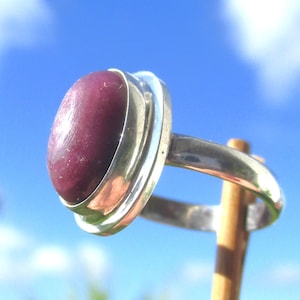 Könnte beinhalten: Ein Silberring mit einem großen, ovalen, tiefroten Edelstein. Der Edelstein ist in einer Lünettenfassung gefasst, und der Ring ist aus poliertem Silber. Der Hintergrund ist ein hellblauer Himmel mit weißen Wolken.
