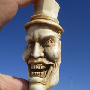 Könnte beinhalten: Eine geschnitzte, cremefarbene Figur eines lächelnden Gesichts mit Zylinder. Das Gesicht hat übertriebene Merkmale, darunter ein breites Grinsen, das Zähne zeigt, eine markante Nase und detaillierte Falten. Der Hintergrund ist ein klarer blauer Himmel.