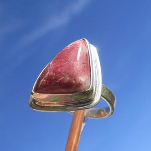 Könnte beinhalten: Ein Silberring mit einem dreieckigen, polierten roten Stein mit weißen Adern, der in einer glatten Metallfassung sitzt. Der Ring ist auf einem dünnen, abgerundeten Silberband montiert und zeigt eine saubere, metallische Oberfläche.