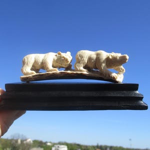 Könnte beinhalten: Eine geschnitzte Skulptur mit zwei cremefarbenen Bären auf einem Holzsockel. Die Bären sind in einer Laufpose dargestellt, mit detailliertem Fell und Gesichtszügen. Die Skulptur steht auf einem schwarzen, rechteckigen Sockel vor blauem Himmel.