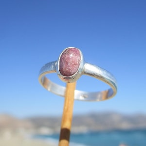 Puede incluir: Un anillo de plata con una gema ovalada de color rosa. El anillo se sostiene contra un cielo azul brillante y un fondo borroso del océano. La gema está engastada en un engaste sencillo.