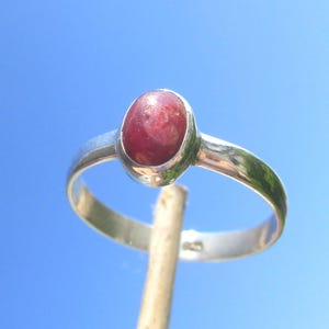 Könnte beinhalten: Ein Silberring mit einem glatten, ovalen, tiefroten Cabochon-Edelstein in einer einfachen Fassung. Die Ringschiene besteht aus poliertem Silber mit einem sichtbaren Stempel auf der Innenseite und einer glänzenden metallischen Oberfläche.