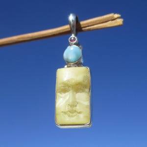 Könnte beinhalten: Ein gelber, geschnitzter Gesichtsanhänger mit einem blauen und einem roten Edelstein. Der Anhänger hängt an einer silbernen Kette.