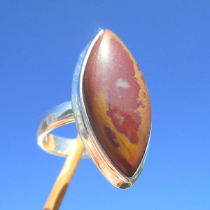 Könnte beinhalten: Ein Silberring mit einem großen, ovalen Stein. Der Stein weist eine Mischung aus Farben auf, darunter Rot, Braun und Gelb. Der Ring ist vor einem leuchtend blauen Himmel platziert.