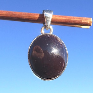 Può includere: Un ciondolo di forma ovale con una pietra liscia color marrone scuro incastonata in una cornice di metallo color argento lucido. Il ciondolo presenta un gancio argentato sulla parte superiore ed è appeso a un bastoncino di legno orizzontale.