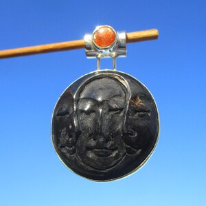 May include: A circular silver pendant featuring a carved black face design. The pendant has a silver bail with a small orange gemstone. The background is a clear blue sky.