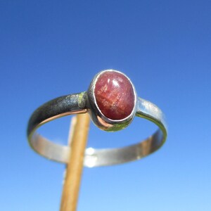 Può includere: Un anello d'argento con una pietra preziosa ovale, marrone rossiccio. La fascia dell'anello è in argento lucido. La pietra preziosa è incastonata in una lunetta d'argento. L'anello è esposto contro un cielo blu brillante.
