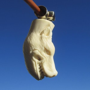 Puede incluir: Un colgante tallado de color crema con forma de cabeza de lobo, con textura de pelaje detallada y la boca abierta, suspendido de una anilla de metal plateado sujeta a un fino palo de madera marrón sobre un fondo de cielo azul claro.