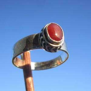 Peut inclure: Une bague en argent ornée d'une pierre précieuse ovale, rouge-orangé. La bague présente un motif décoratif autour de la pierre et un large anneau. Le fond est un ciel bleu clair et lumineux.