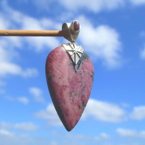 Op de afbeelding: Een traanvormige rhodoniet hanger met een gevlekt roze en zwart stenen oppervlak. De hanger is voorzien van een zilverkleurig metalen oogje met een stervormige gravure en is versierd met een kleine, ronde, donkerrode edelsteen.